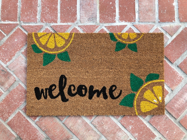 K&ouml;nnte beinhalten: Rechteckige Fu&szlig;matte aus braunem Kokosfasermaterial. Der Schriftzug "welcome" ist in schwarzer Schrift in der Mitte zu sehen. Die Matte ist mit gelben und gr&uuml;nen Zitronenscheiben-Designs in jeder Ecke verziert.