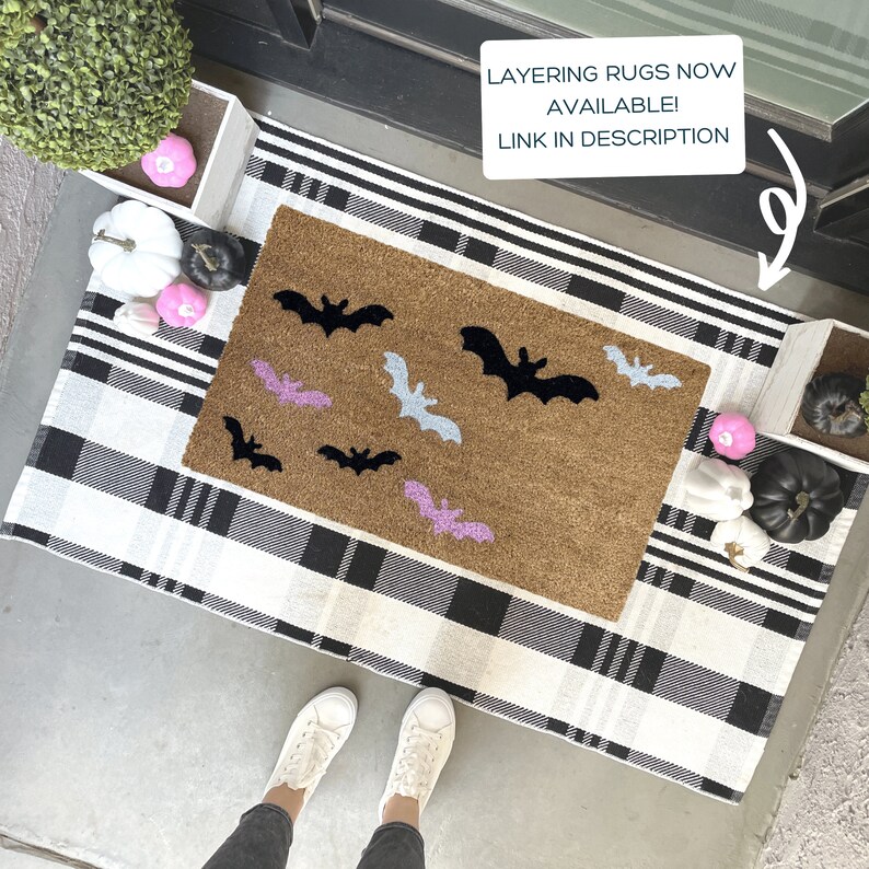 May include: A brown doormat with black and white bats on it. The doormat is on a black and white checkered rug. The text "LAYERING RUGS NOW AVAILABLE! LINK IN DESCRIPTION" is on a white sign above the doormat.