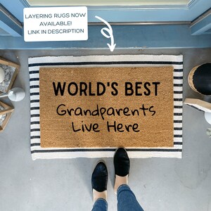 May include: A brown coir doormat with black and white striped border. The doormat has the text "WORLD'S BEST Grandparents Live Here" written in black.