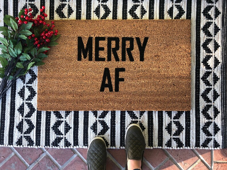 May include: A brown coir doormat with the text "MERRY AF" in black lettering. The doormat is on a black and white patterned rug.