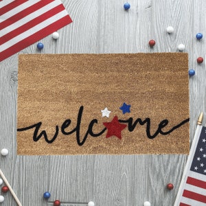 May include: A brown welcome mat with the word "welcome" in black script. The word is decorated with red, white, and blue stars.