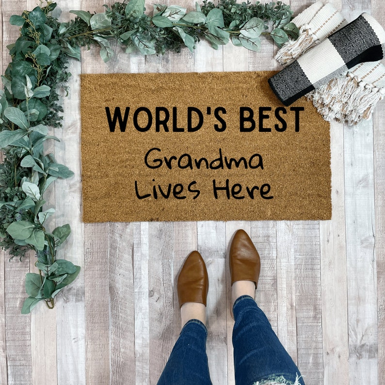 May include: A brown doormat with the text "WORLD'S BEST Grandma Lives Here" in black lettering.