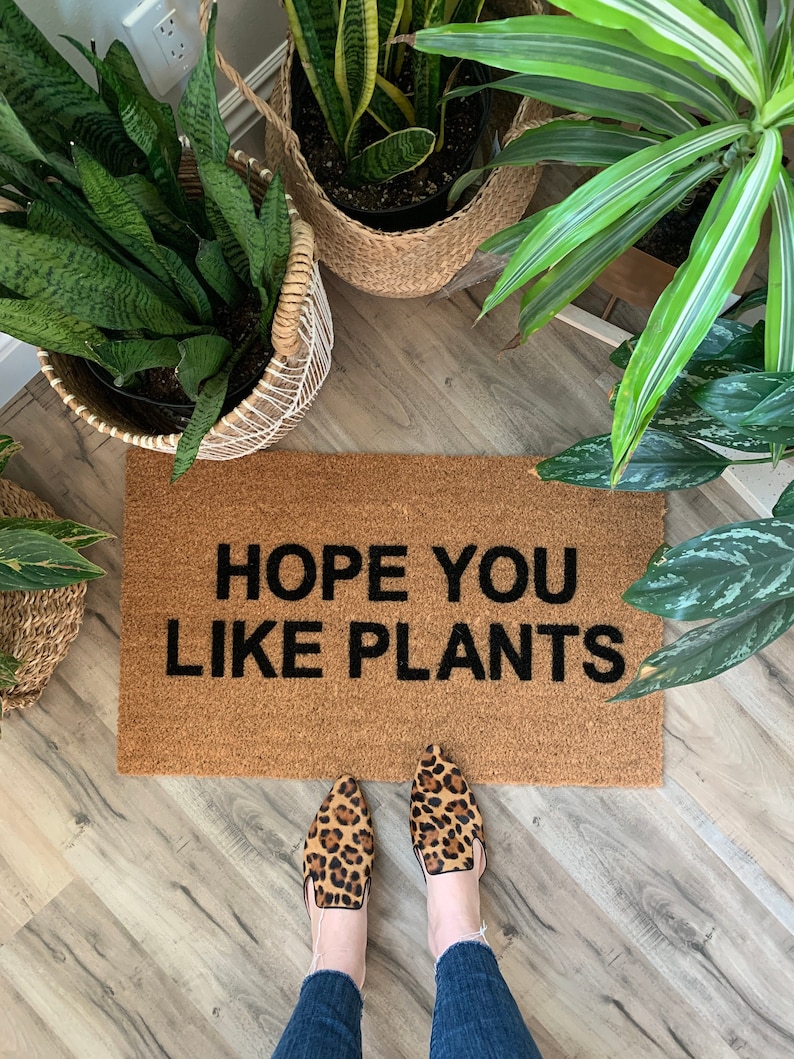 K&ouml;nnte beinhalten: Eine braune Fu&szlig;matte mit dem Text "HOPE YOU LIKE PLANTS" in schwarzen Buchstaben. Ein Paar Leopardenmuster-Schuhe befinden sich auf der Fu&szlig;matte.