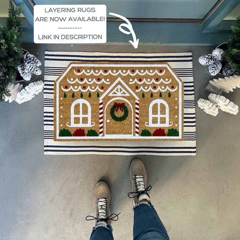 May include: A brown and white gingerbread house doormat with blue and white stripes. The doormat is on a gray concrete floor. The text "LAYERING RUGS ARE NOW AVAILABLE! LINK IN DESCRIPTION" is visible in the image.