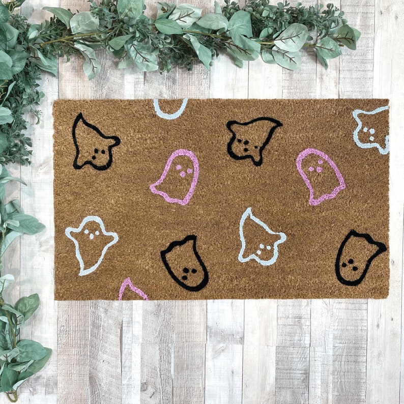 Pode incluir: Um tapete de porta marrom com tema de Halloween com v&aacute;rios desenhos de fantasmas em rosa, azul e preto.