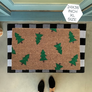 May include: A brown doormat with a black and white checkered border. The doormat has a pattern of green Christmas trees. The text "24X36 INCH XL SIZE" is printed on a white hexagonal sticker.
