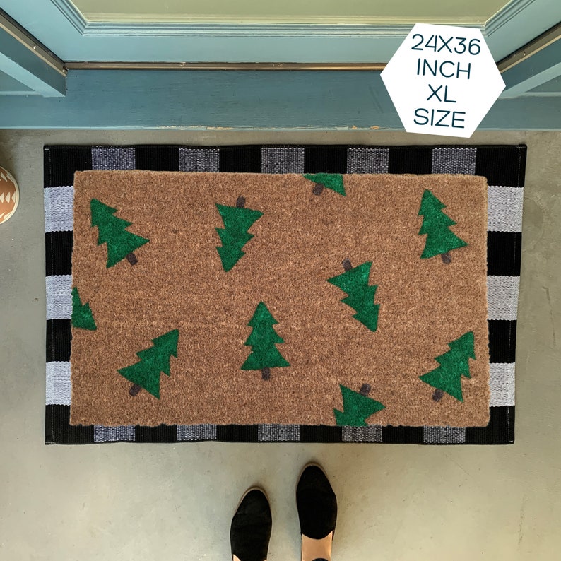 May include: A brown doormat with a black and white checkered border. The doormat has a pattern of green Christmas trees. The text "24X36 INCH XL SIZE" is printed on a white hexagonal sticker.