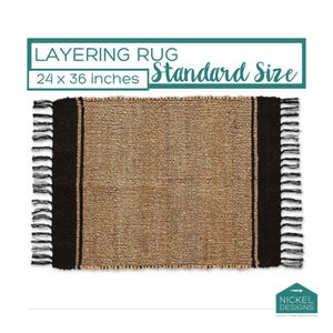 May include: A rectangular layering rug measuring 24 x 36 inches with a woven tan center and solid black side panels featuring fringed edges. The text LAYERING RUG 24 x 36 inches Standard Size and the Nickel Designs logo are visible.