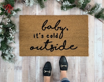 May include: A brown doormat with the text "baby, it's cold outside" in black cursive lettering. A pair of black leather loafers with gold buckles are on a light wood floor.