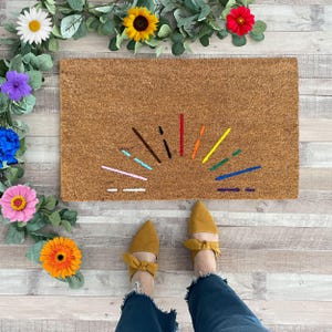 May include: A rectangular brown coir doormat with a colorful embroidered sunburst design. The design includes lines in various colors, including red, orange, yellow, green, blue, and purple. Yellow shoes with bow details are visible.