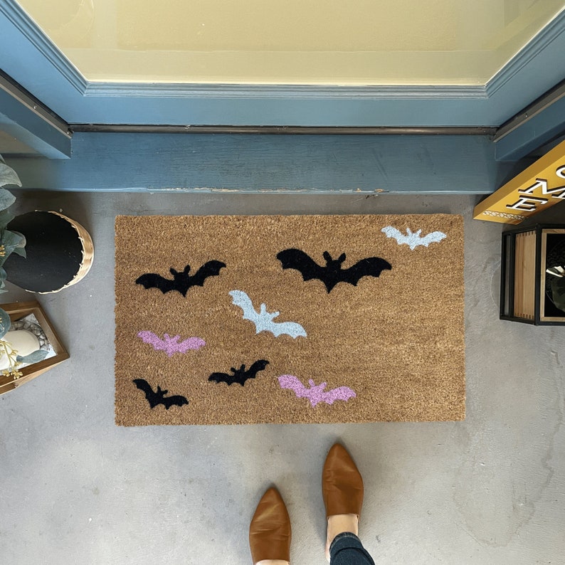 May include: A brown coir doormat with a Halloween theme featuring black and light blue bats and pink bats.