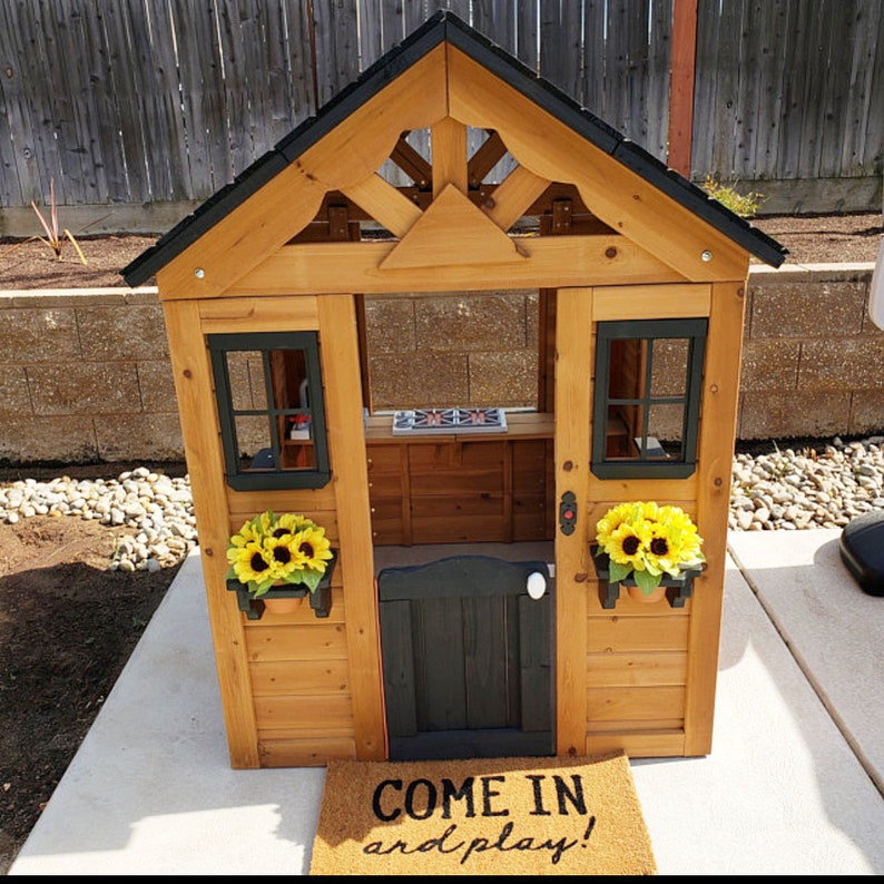 May include: A wooden playhouse with a black roof and a black door. The playhouse has two windows with black frames and a small porch with a black railing. There are two potted sunflowers on either side of the door. The playhouse has a sign that says "Come in and play!" on the ground in front of it.