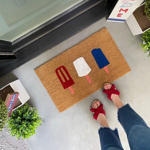 May include: A brown doormat with four popsicle shapes in red, white, and blue. The popsicles are arranged in a row, with the red popsicle on the left and the blue popsicle on the right. The doormat is on a concrete surface.