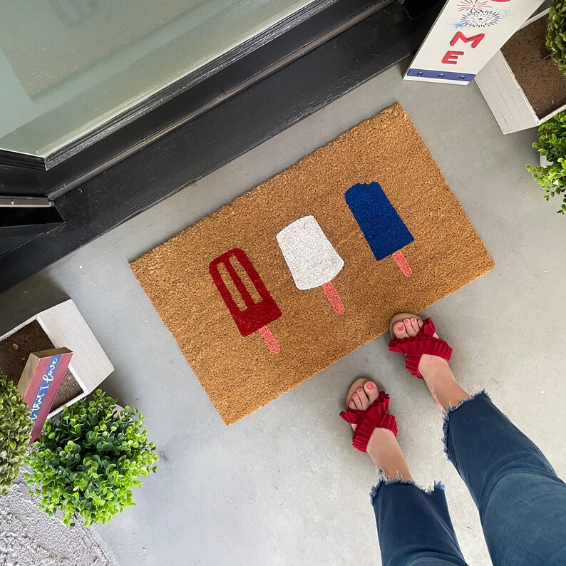 May include: A brown doormat with four popsicle shapes in red, white, and blue. The popsicles are arranged in a row, with the red popsicle on the left and the blue popsicle on the right. The doormat is on a concrete surface.