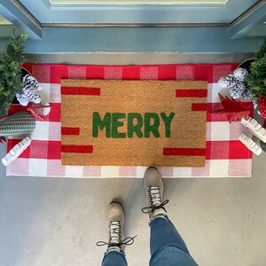 May include: A brown doormat with the word "MERRY" in green letters on a white and red checkered background. A pair of brown boots with white laces are on the mat.