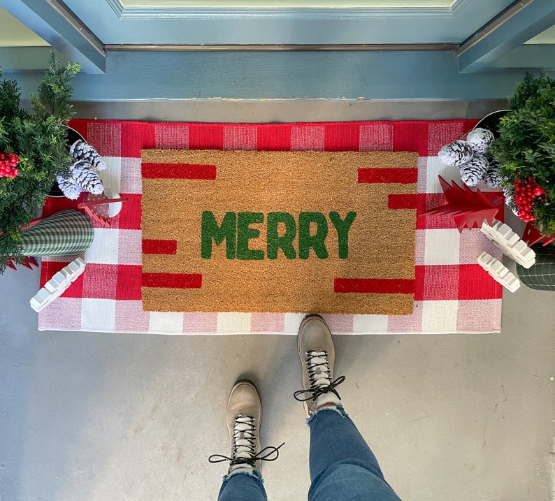 May include: A brown doormat with the word "MERRY" in green letters on a white and red checkered background. A pair of brown boots with white laces are on the mat.