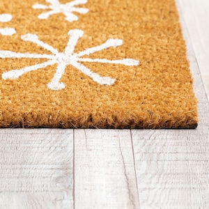 May include: A brown coir doormat with white snowflake designs. The doormat is on a light wood floor.