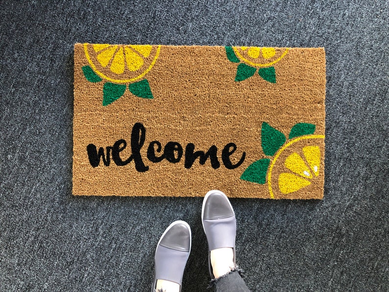 K&ouml;nnte beinhalten: Rechteckige Fu&szlig;matte aus braunem Kokosfasermaterial mit dem schwarzen Schriftzug "welcome". Zitronenscheiben-Designs in Gelb und Gr&uuml;n an den Ecken. Eine Person steht auf der Matte.