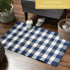May include: A rectangular blue and white checkered rug made of 100% cotton. The rug is placed on a wooden floor, with a pair of gray slippers and a wicker basket nearby. The text "100% Cotton Perfect or indoors too!" is visible.