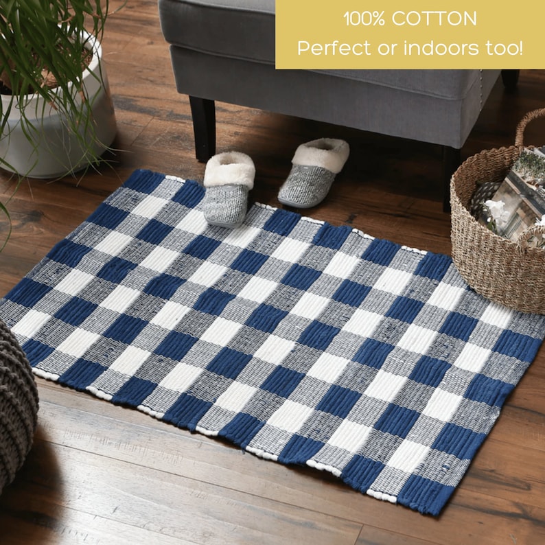 May include: A rectangular blue and white checkered rug made of 100% cotton. The rug is placed on a wooden floor, with a pair of gray slippers and a wicker basket nearby. The text "100% Cotton Perfect or indoors too!" is visible.