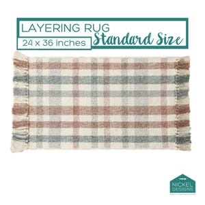 May include: A rectangular layering rug measuring 24 x 36 inches with a cream base and a plaid pattern in muted terracotta and sage green. The rug features fringed edges on the left and right sides. Text above reads: LAYERING RUG Standard Size.