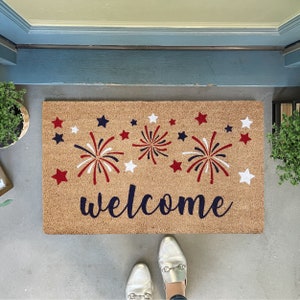 May include: A brown coir doormat with the word "welcome" in black script. The doormat is decorated with red, white, and blue fireworks and stars.