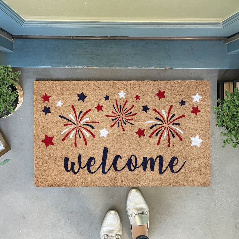 May include: A brown coir doormat with the word "welcome" in black script. The doormat is decorated with red, white, and blue fireworks and stars.