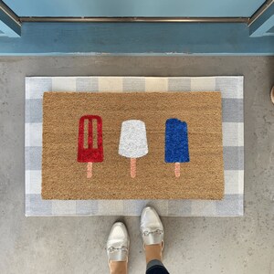 May include: A brown doormat with a white and grey checkered border. The doormat has three popsicle designs on it: one red, one white, and one blue.
