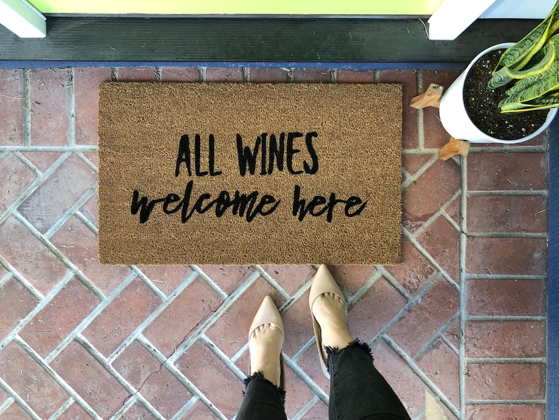 May include: A rectangular doormat made of natural fibers with the text "ALL WINES welcome here" in black script. The mat is on a brick surface, with a pair of beige shoes and a potted plant visible.