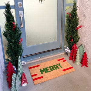 May include: A brown doormat with the word "Merry" in green letters and red stripes. The doormat is on a wooden floor in front of a door with a wreath hanging on it. There are small artificial Christmas trees in pots on either side of the door.