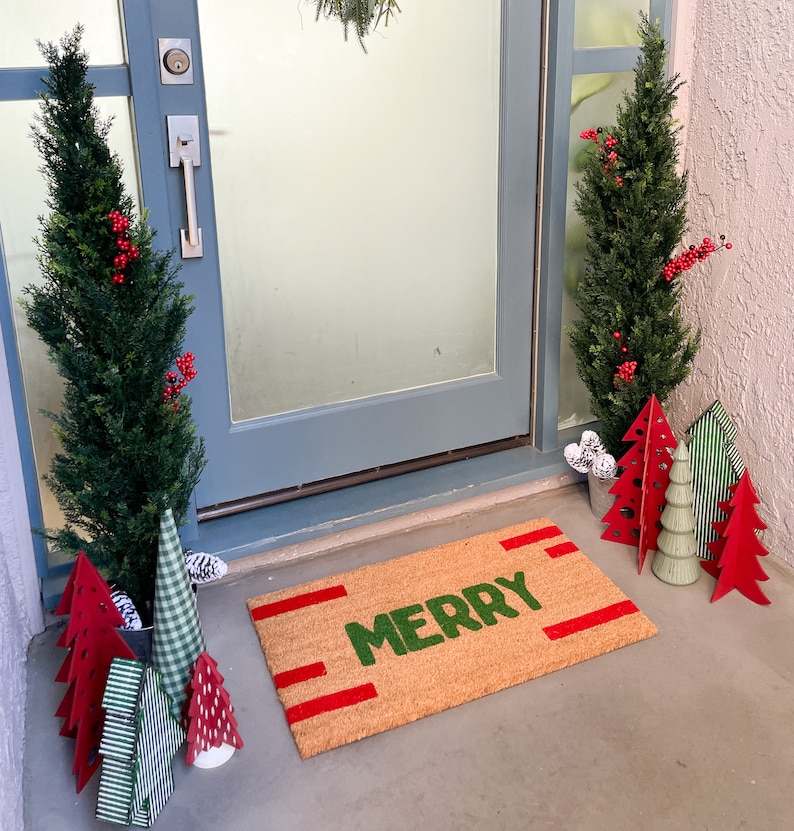 May include: A brown doormat with the word "Merry" in green letters and red stripes. The doormat is on a wooden floor in front of a door with a wreath hanging on it. There are small artificial Christmas trees in pots on either side of the door.