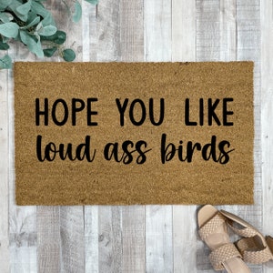 May include: A brown coir doormat with the text "HOPE YOU LIKE LOUD ASS BIRDS" in black lettering.