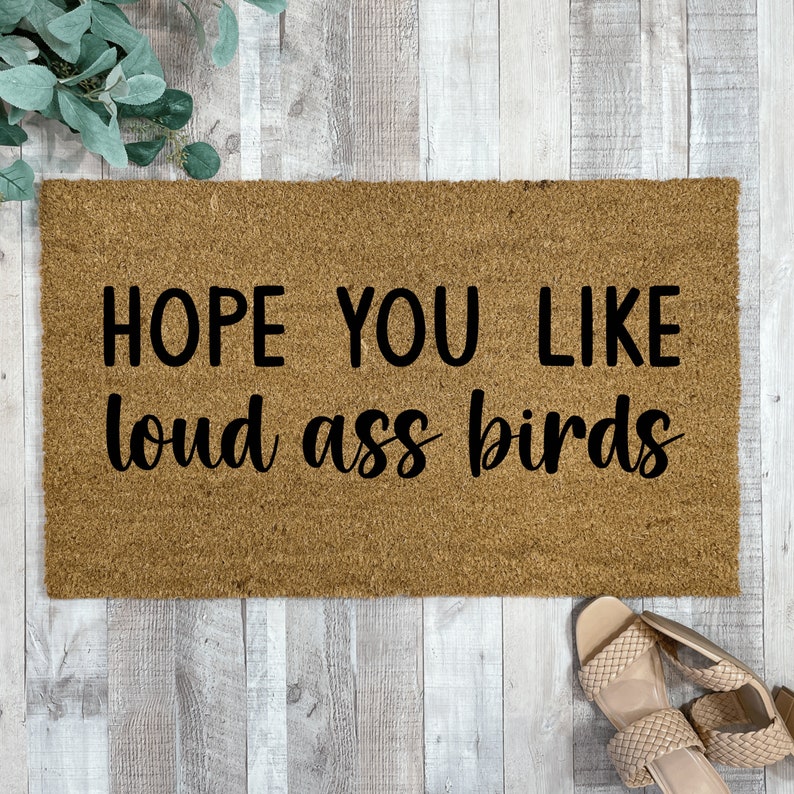 May include: A brown coir doormat with the text "HOPE YOU LIKE LOUD ASS BIRDS" in black lettering.