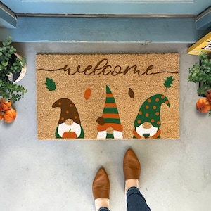 May include: A rectangular coir doormat with the word "welcome" in a cursive font. The mat features three gnome figures with pointed hats in brown, green, and orange, with fall leaf accents. The mat is placed in front of a blue door.
