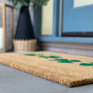 May include: A brown coir doormat with a green leaf design. The doormat is placed on a concrete surface in front of a blue door.
