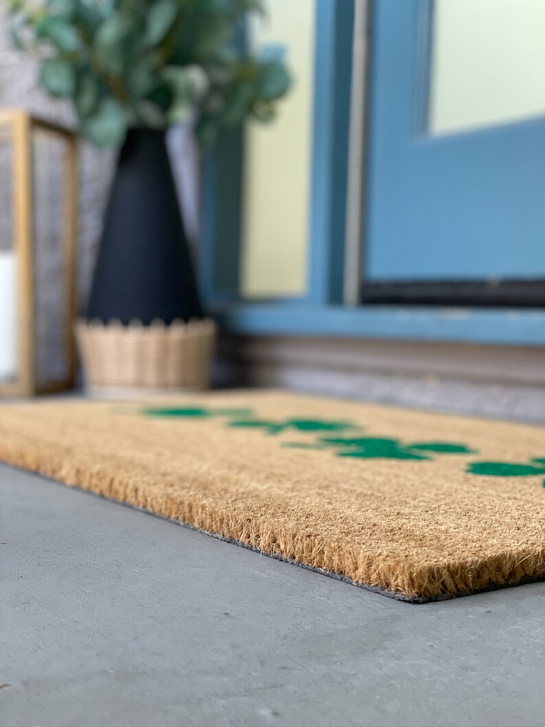 May include: A brown coir doormat with a green leaf design. The doormat is placed on a concrete surface in front of a blue door.