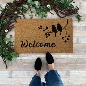 Puede incluir: Una alfombra de coco marr&oacute;n con la palabra "Welcome" y una silueta de tres p&aacute;jaros en una rama con hojas.