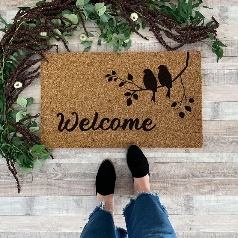 Puede incluir: Una alfombra de coco marr&oacute;n con la palabra "Welcome" y una silueta de tres p&aacute;jaros en una rama con hojas.
