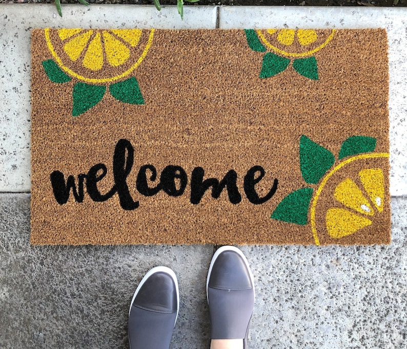 K&ouml;nnte beinhalten: Rechteckige Fu&szlig;matte aus brauner Kokosfaser. Das Wort "welcome" ist in schwarzer Schrift geschrieben. Gelbe Zitronenscheiben mit gr&uuml;nen Bl&auml;ttern befinden sich in jeder Ecke. Graue Slipper sind unten.