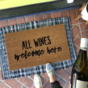May include: A brown doormat with the text "ALL WINES welcome here" sits on a blue and white patterned rug. A person is wearing silver loafers and black jeans. A bottle of white wine is held in the person's hand.