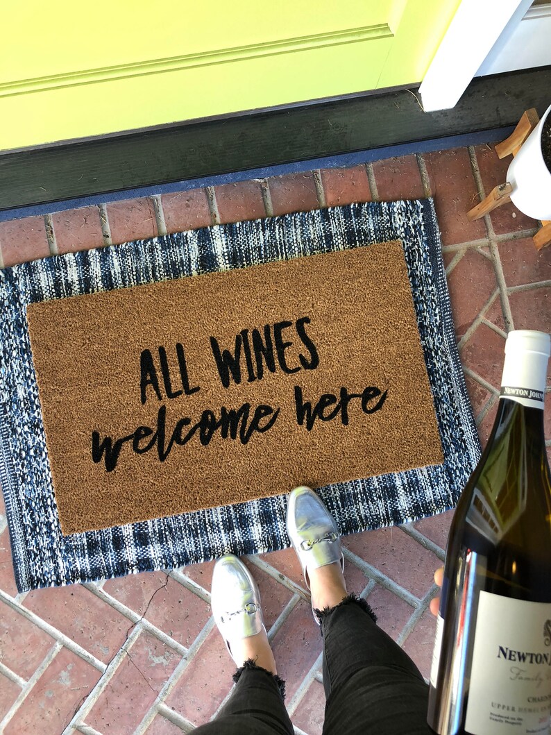 May include: A brown doormat with the text "ALL WINES welcome here" sits on a blue and white patterned rug. A person is wearing silver loafers and black jeans. A bottle of white wine is held in the person's hand.