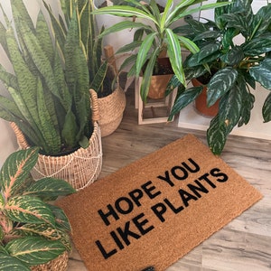 K&ouml;nnte beinhalten: Eine braune Fu&szlig;matte mit dem Text "HOPE YOU LIKE PLANTS" in schwarzen Buchstaben. Die Fu&szlig;matte liegt auf einem Holzboden mit mehreren Topfpflanzen im Hintergrund.