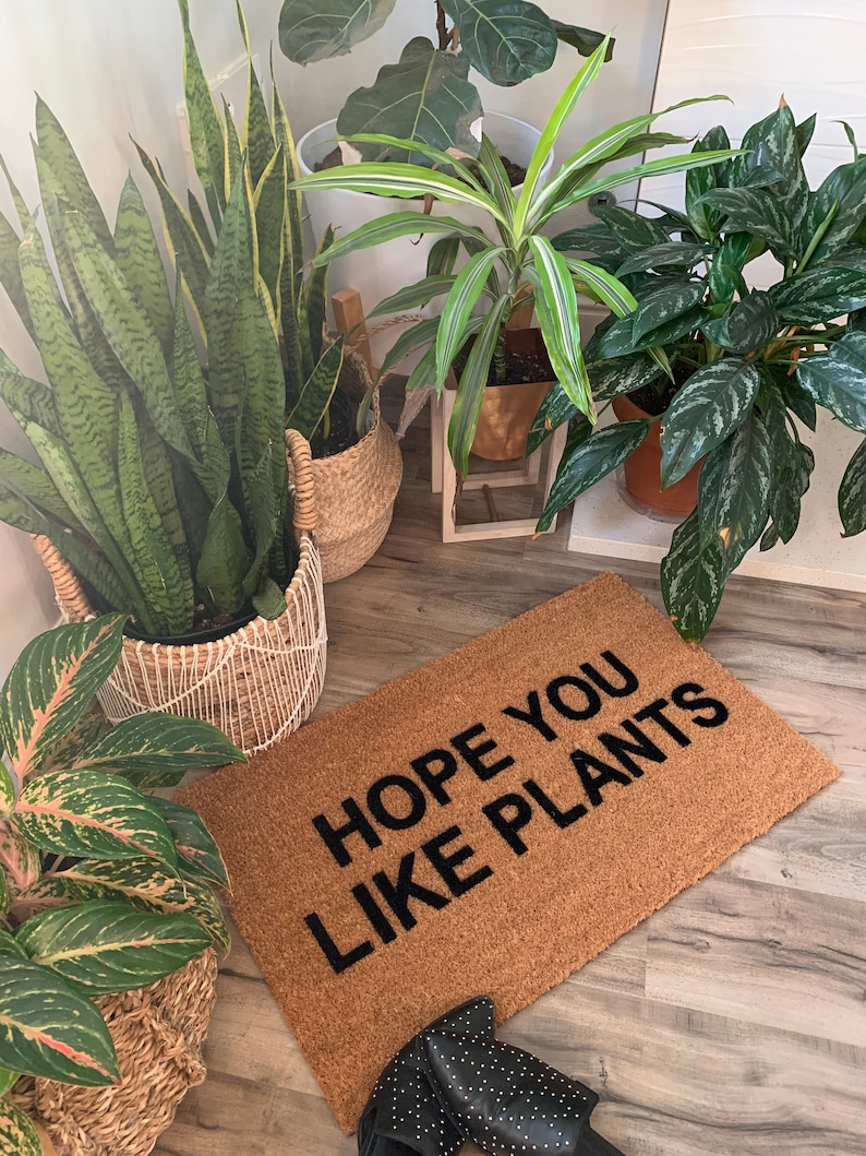 K&ouml;nnte beinhalten: Eine braune Fu&szlig;matte mit dem Text "HOPE YOU LIKE PLANTS" in schwarzen Buchstaben. Die Fu&szlig;matte liegt auf einem Holzboden mit mehreren Topfpflanzen im Hintergrund.