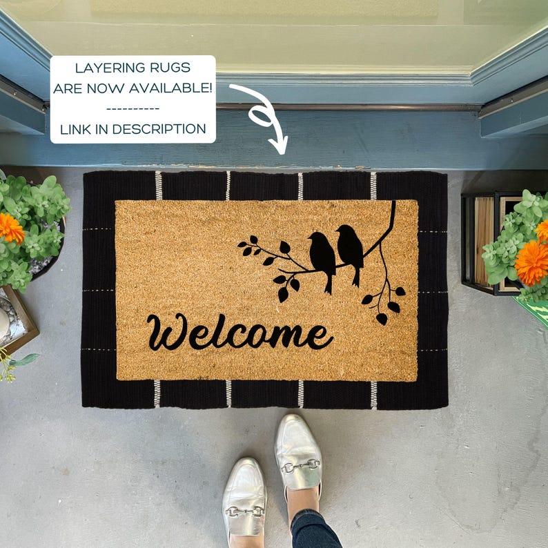 Puede incluir: Una alfombra de bienvenida de fibra de coco marr&oacute;n con ribete negro y rayas blancas. La alfombra tiene una silueta negra de tres p&aacute;jaros en una rama con la palabra "Bienvenido" en letras cursivas negras.