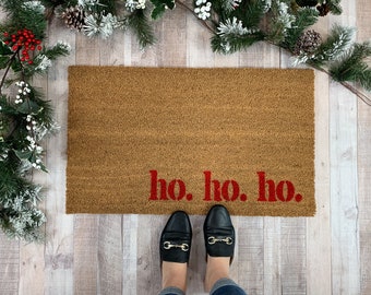 May include: A rectangular tan coir doormat featuring the red text ho. ho. ho. printed in the lower right corner. The mat has a coarse, fibrous texture and is positioned on a light-coloured wooden floor surface with festive greenery nearby.