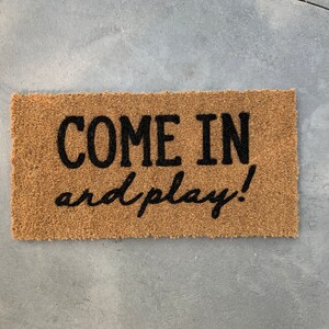 May include: A brown coir doormat with the text "COME IN and play!" in black lettering.
