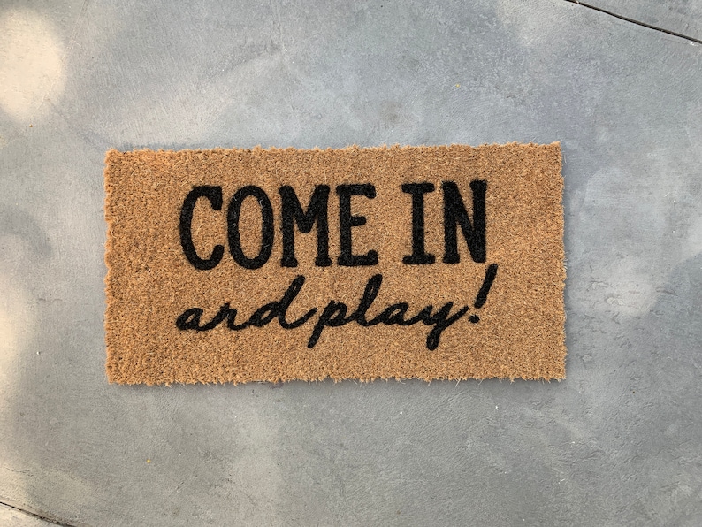 May include: A brown coir doormat with the text "COME IN and play!" in black lettering.