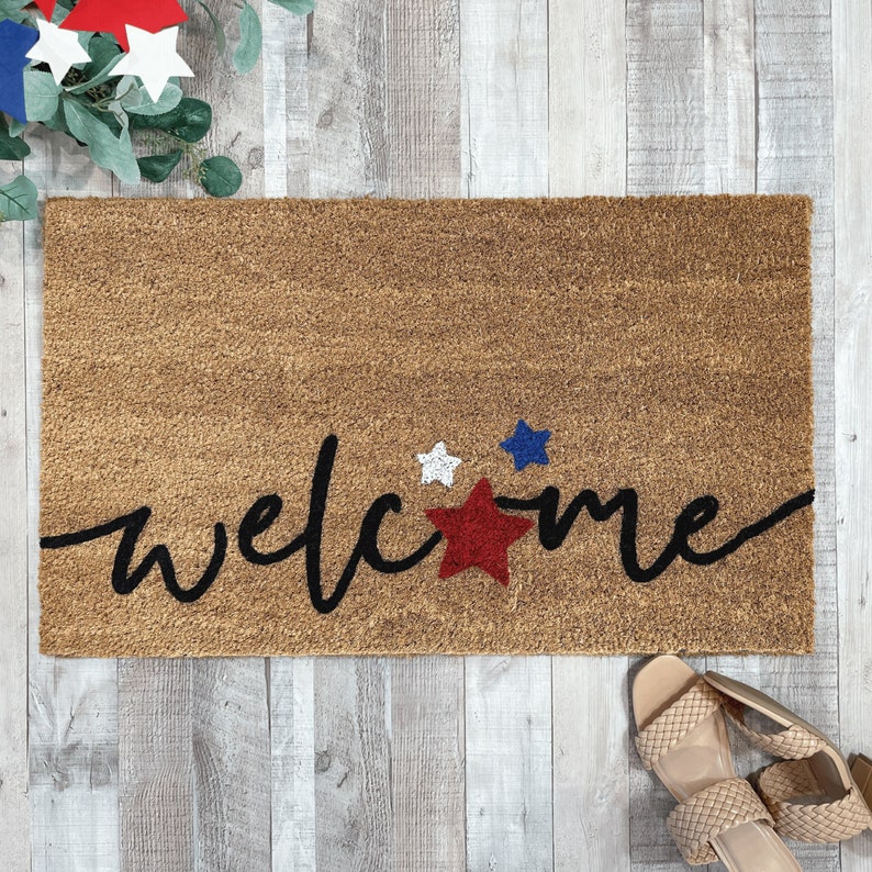 May include: A brown coir doormat with the word "welcome" written in black cursive script. There are three red, white, and blue stars in the center of the mat.