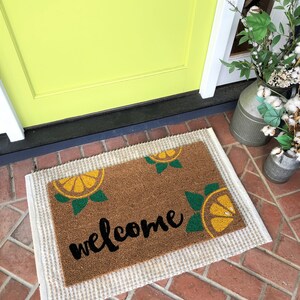 K&ouml;nnte beinhalten: Eine rechteckige Fu&szlig;matte mit einer braunen Kokosoberfl&auml;che und einem wei&szlig;en Rand. Die Matte zeigt das Wort "welcome" in schwarzer Schrift sowie gelbe und gr&uuml;ne Zitronenscheiben-Designs. Die Matte liegt vor einer limettengr&uuml;nen T&uuml;r.