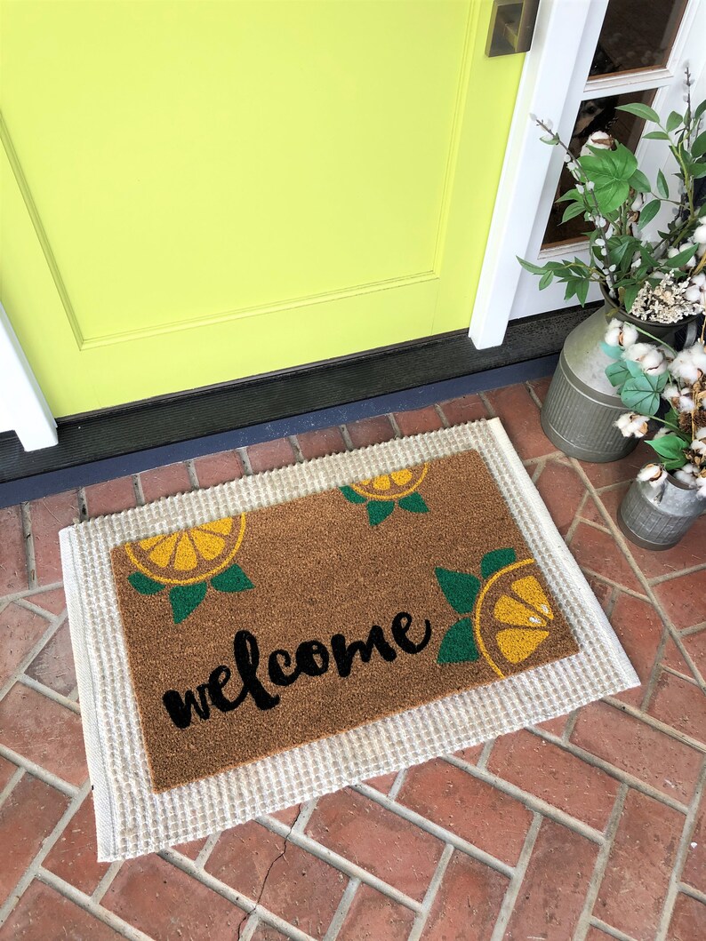 K&ouml;nnte beinhalten: Eine rechteckige Fu&szlig;matte mit einer braunen Kokosoberfl&auml;che und einem wei&szlig;en Rand. Die Matte zeigt das Wort "welcome" in schwarzer Schrift sowie gelbe und gr&uuml;ne Zitronenscheiben-Designs. Die Matte liegt vor einer limettengr&uuml;nen T&uuml;r.